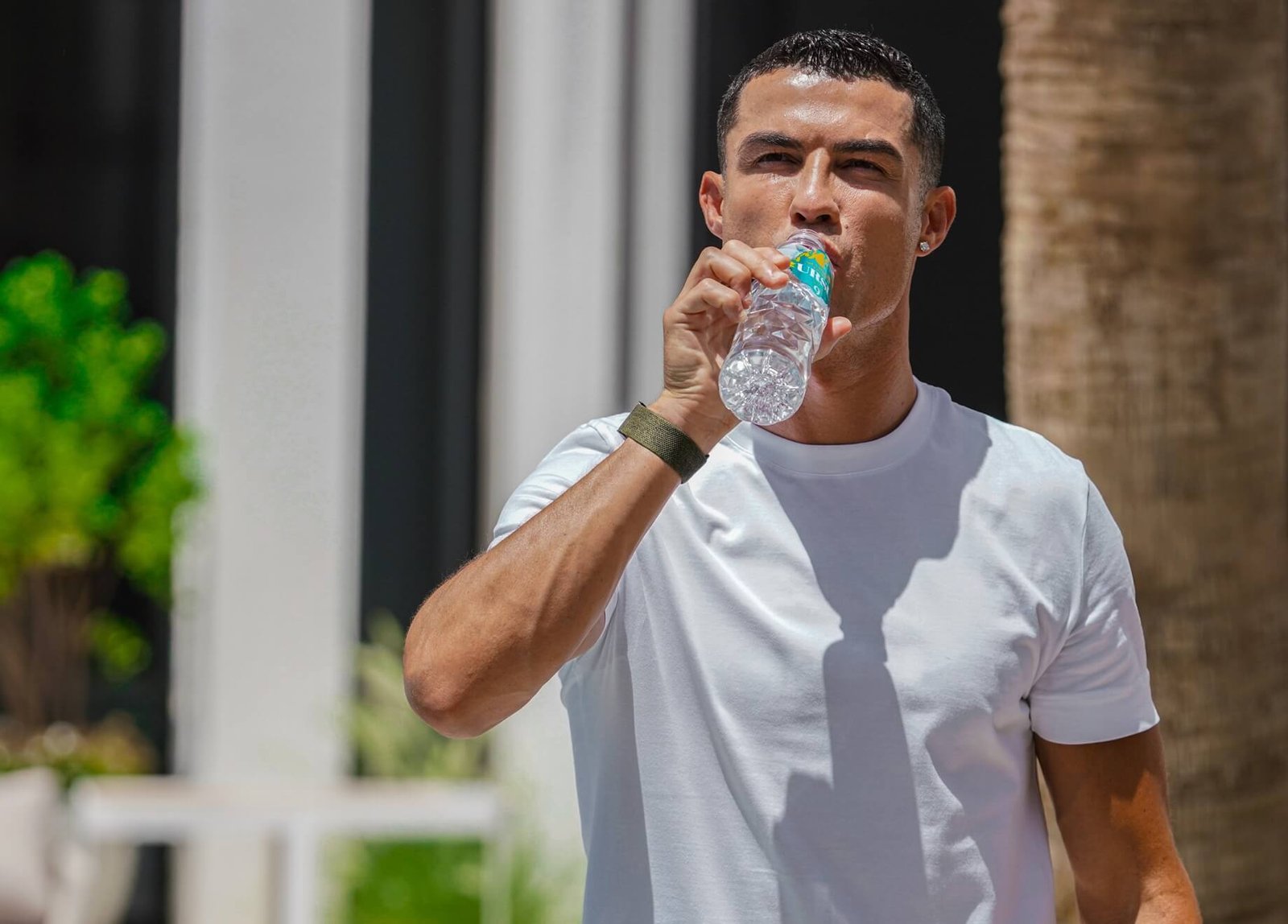 Mann in weißem T-Shirt trinkt Wasser aus einer Flasche der Marke Hotblood. Im Hintergrund sind Pflanzen sichtbar.